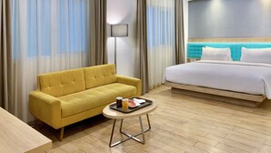 Une chambre d’hôtel avec un canapé jaune, un lit doté d’une tête de lit bleue, une petite table ronde sur laquelle se trouve un plateau de nourriture, et des rideaux légers.