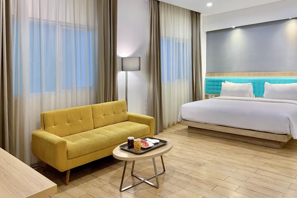 Une chambre d’hôtel avec un canapé jaune, un lit doté d’une tête de lit bleue, une petite table ronde sur laquelle se trouve un plateau de nourriture, et des rideaux légers.