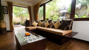 A modern living room with a wooden floor, a sofa with cushions, a coffee table, and a view of a garden and traditional architecture outside. - Villa Nesanur (Denpasar)