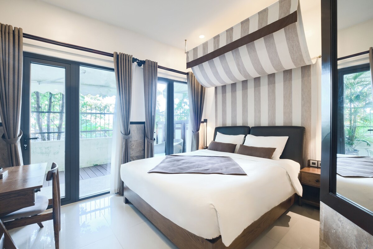 A modern hotel room with a large bed, a wooden desk, and a balcony with curtains.