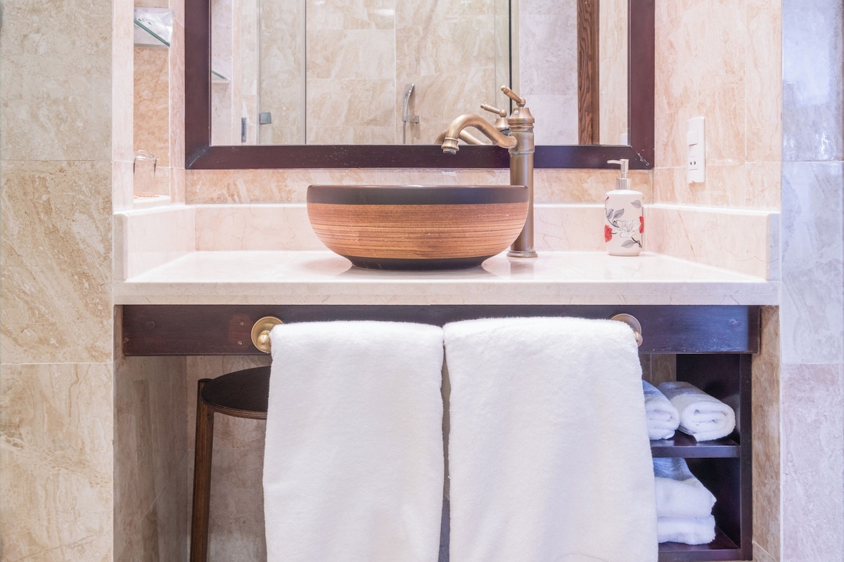 A bathroom with a marble sink, a wooden bowl basin, a brass faucet, and a towel rack with towels.