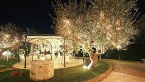 Un espace extérieur aménagé en patio, avec une fontaine en pierre, des tables et des chaises, entouré d’arbres et comprenant un coin salon abrité.