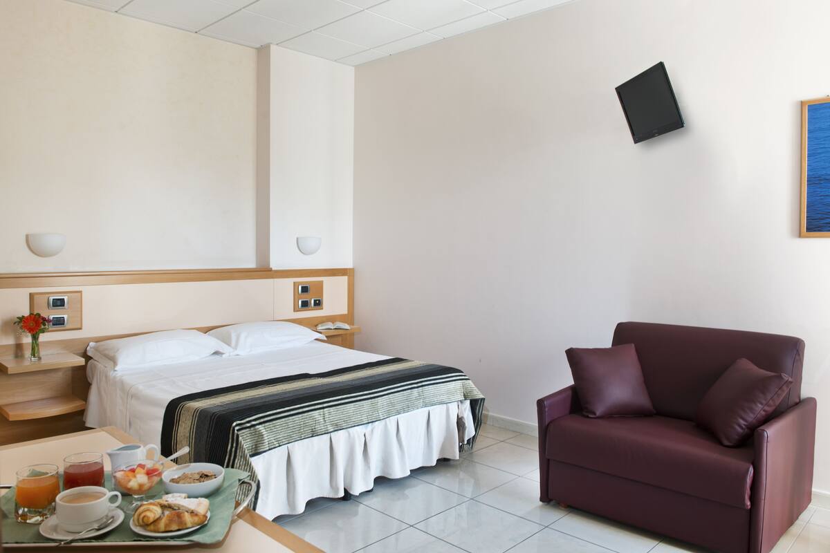 Habitación de hotel con cama, mesitas de noche, una mesa de centro con alimentos para el desayuno, un sofá y un televisor instalado en la pared.