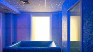 Une pièce avec une baignoire carrelée de carreaux bleus, un banc en bois et une porte donnant sur un espace lumineux.