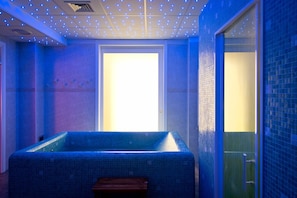 A room with a blue-tiled bathtub, a wooden bench, and a door leading to a bright area. - Park Hotel La Grave (Castellana Grotte)