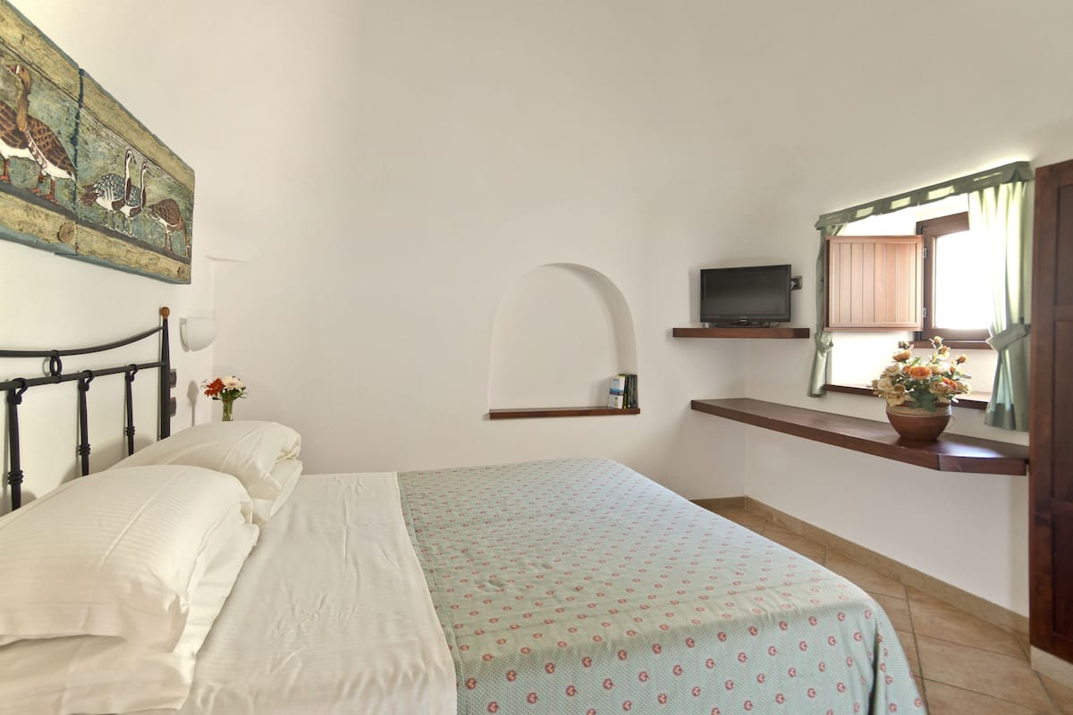 Un dormitorio con una cama, televisión, estante con libros, ventana con cortinas y un espejo adosado a la pared.