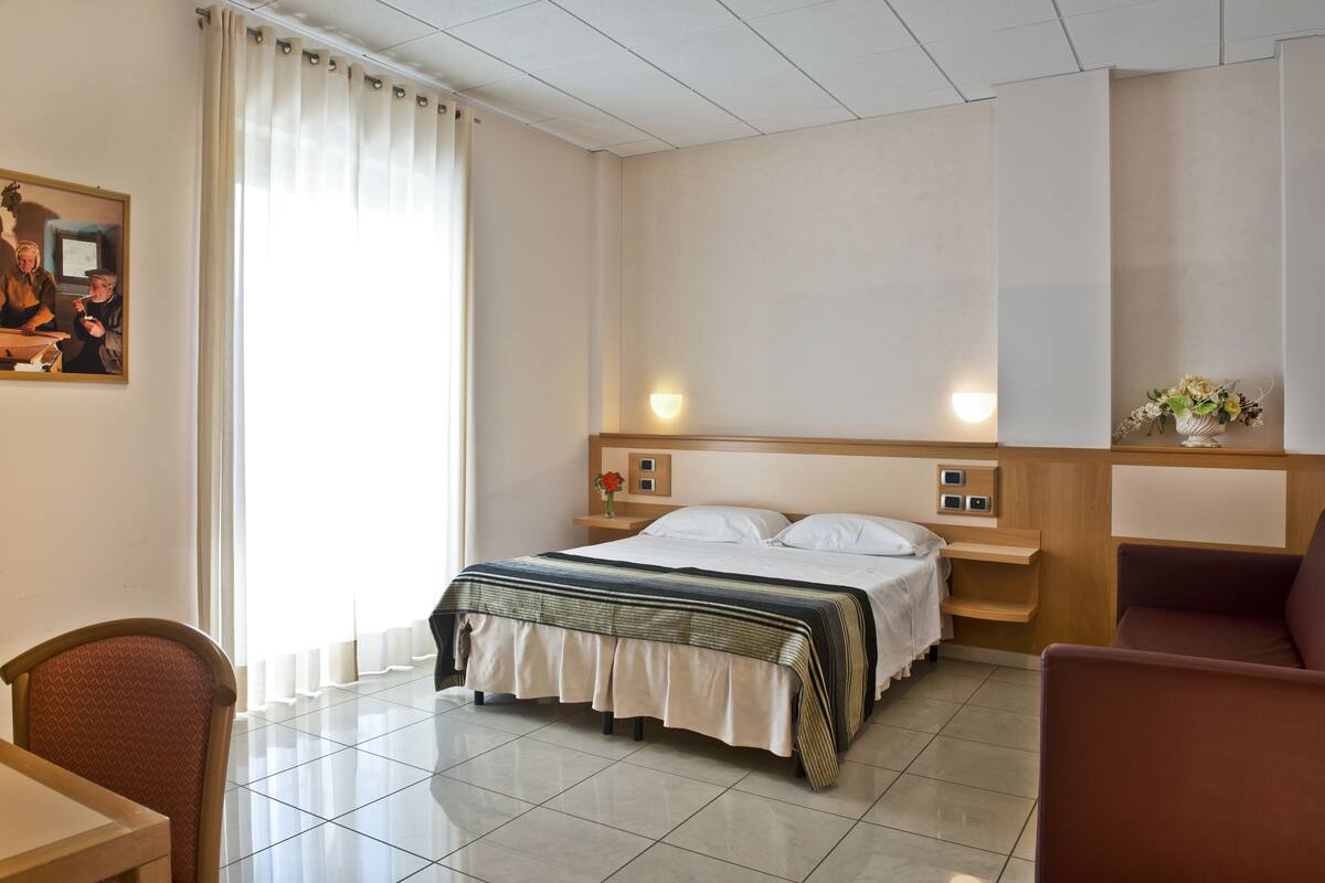 Habitación de hotel con cama, escritorio, silla, mesita de noche y estante fijado a la pared.