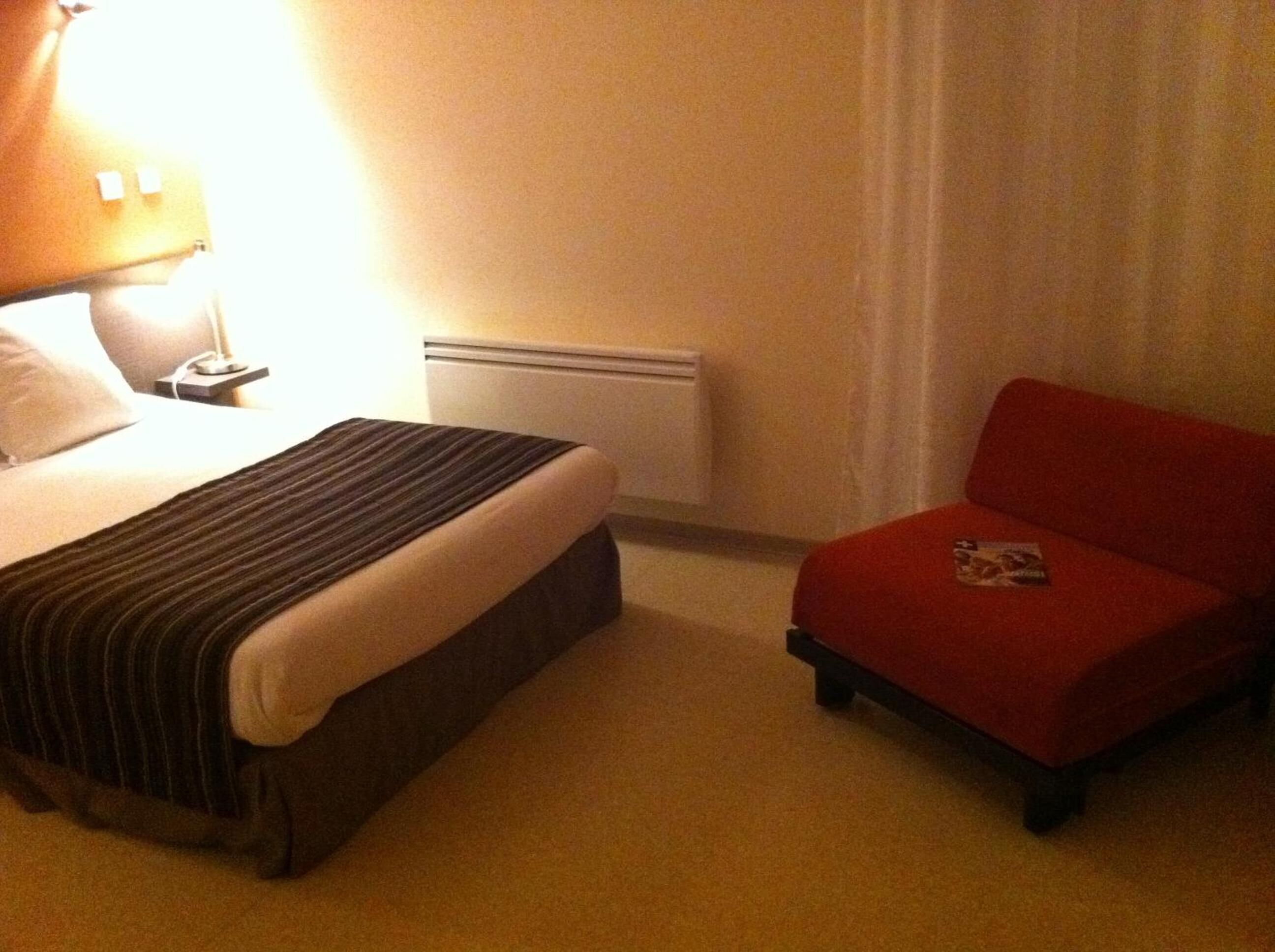 Una habitación de hotel con cama, mesita de noche, una silla roja y ventana con cortinas.