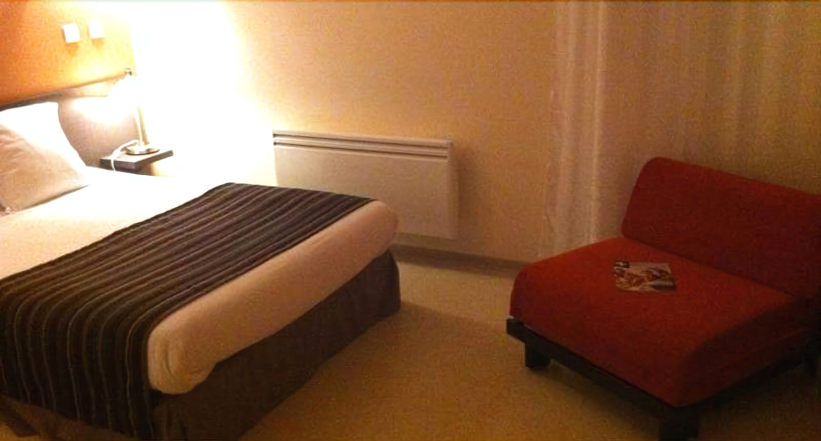 A hotel room with a bed, a nightstand with a lamp, a red chair, and a radiator.