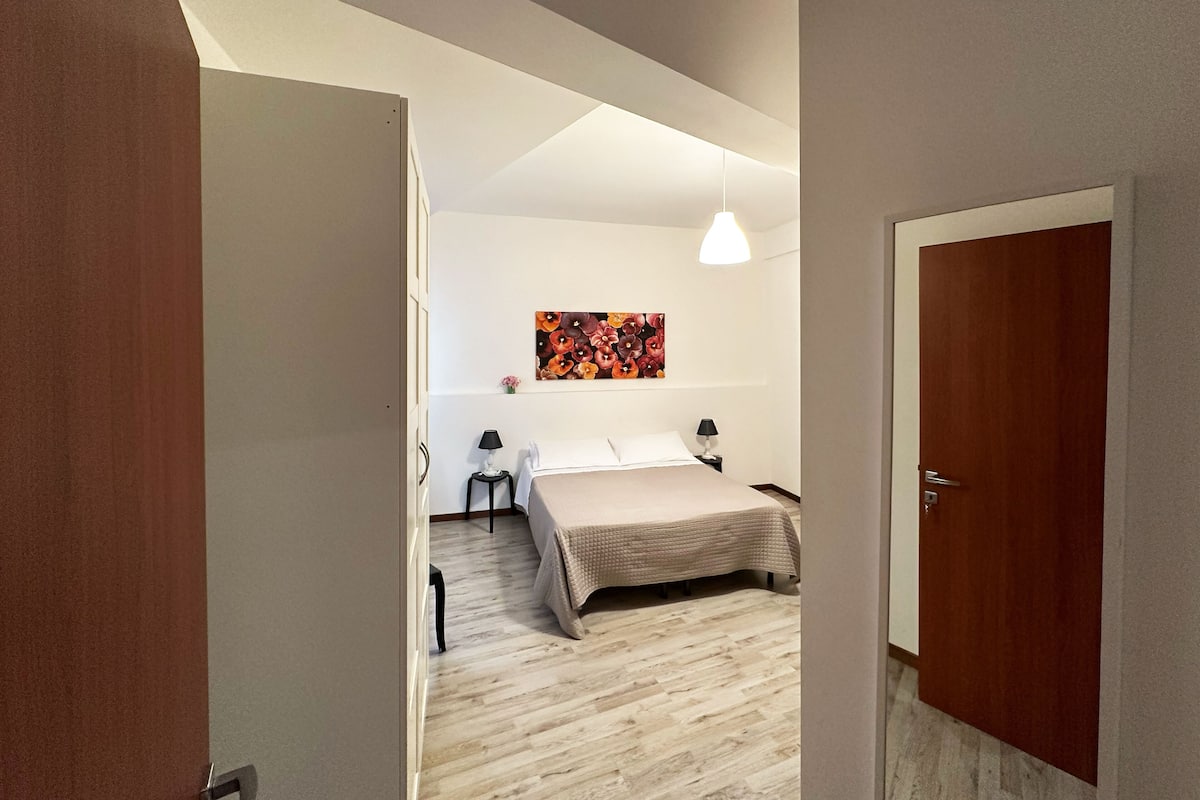 A bedroom with a bed, two bedside tables, a hanging light, and a wall-mounted artwork.
