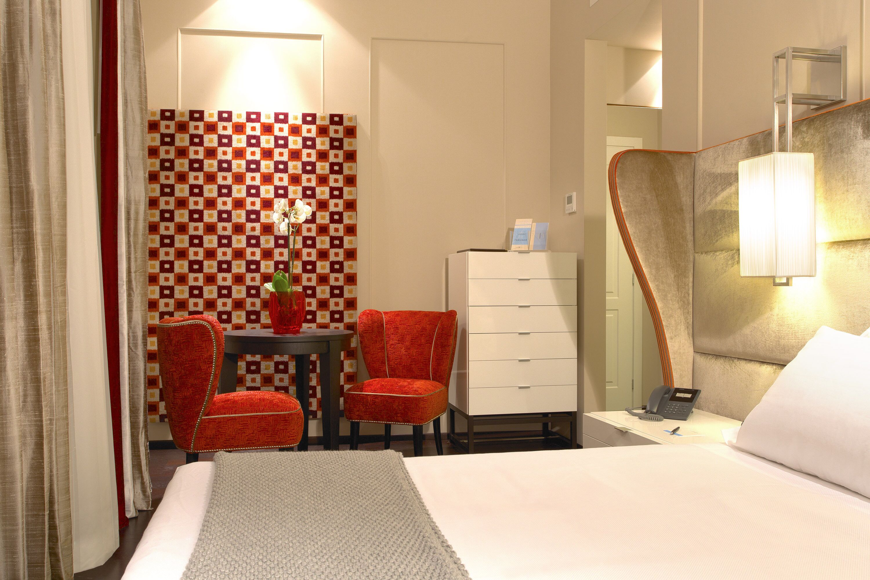 A hotel room with a bed, a desk with a telephone, two red chairs, a small table with a vase, and a patterned wall.