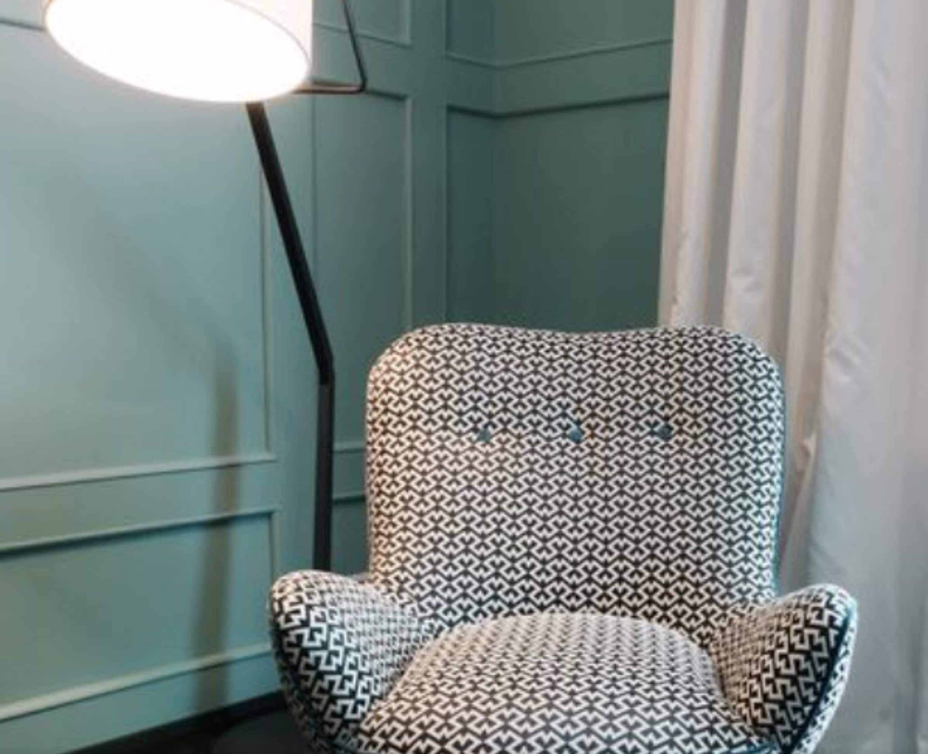 A corner of a room with a patterned armchair, a floor lamp, and a curtain.