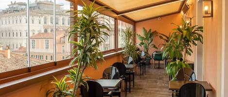 A sunlit indoor space with tables and chairs, large windows, and potted plants.