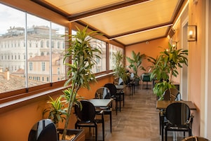 Spazio interno luminoso con tavoli e sedie, ampie finestre e piante in vaso.