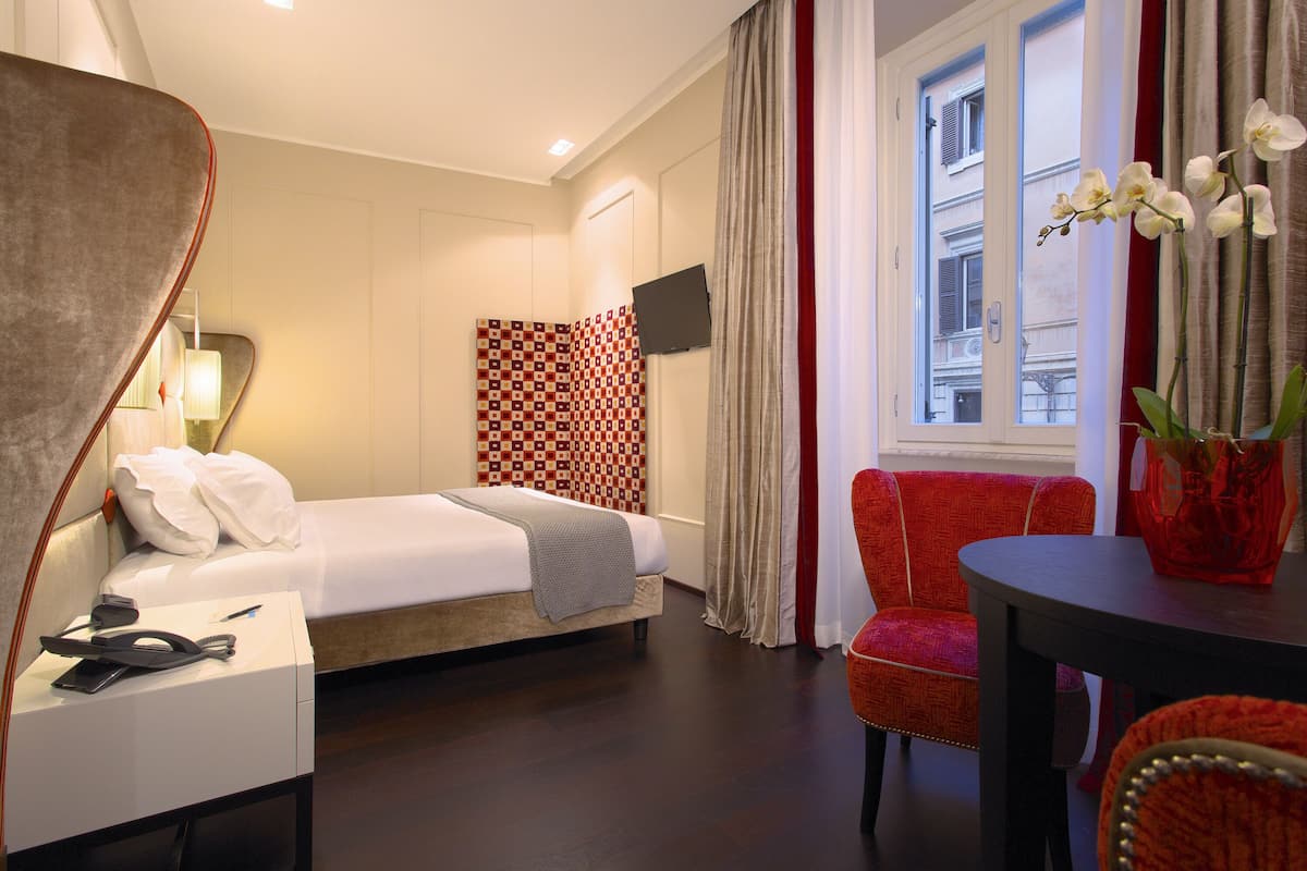 A hotel room with a bed, a desk with a lamp, a chair, a table, and a window with curtains.