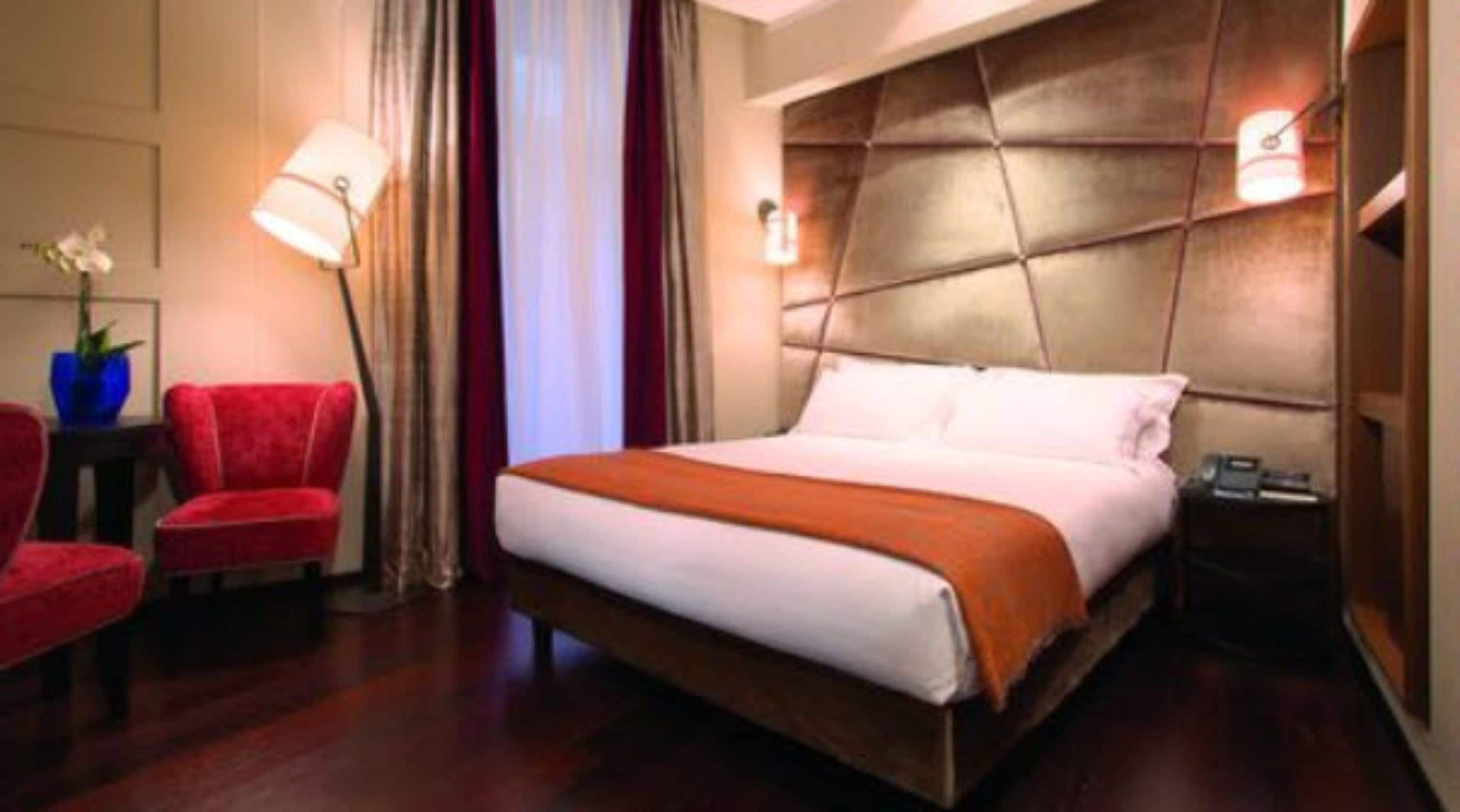 A hotel room with a large bed, a red armchair, a nightstand with a lamp, and a potted plant.