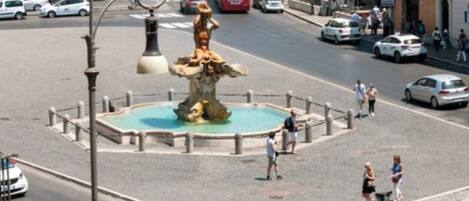 A public square with a central fountain, surrounded by a low fence, and various people walking around.
