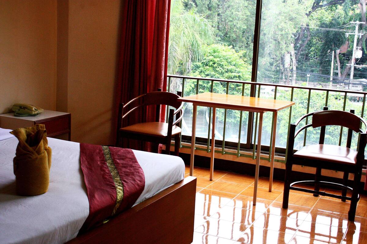A hotel room with a bed, a table, two chairs, a small table with a phone, and a window with a view of greenery.
