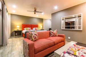 Quarto moderno com sofá vermelho, cama grande com várias almofadas, ventilador de teto e janela com cortinas.