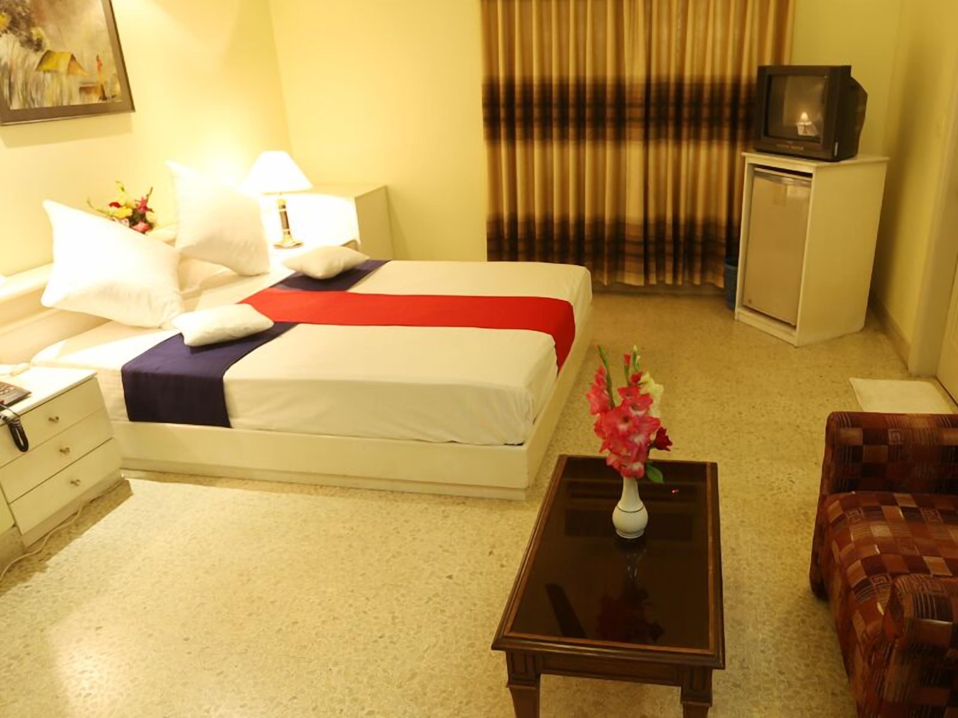 A hotel room with a bed, a nightstand, a desk, a chair, a television, and a vase with flowers.