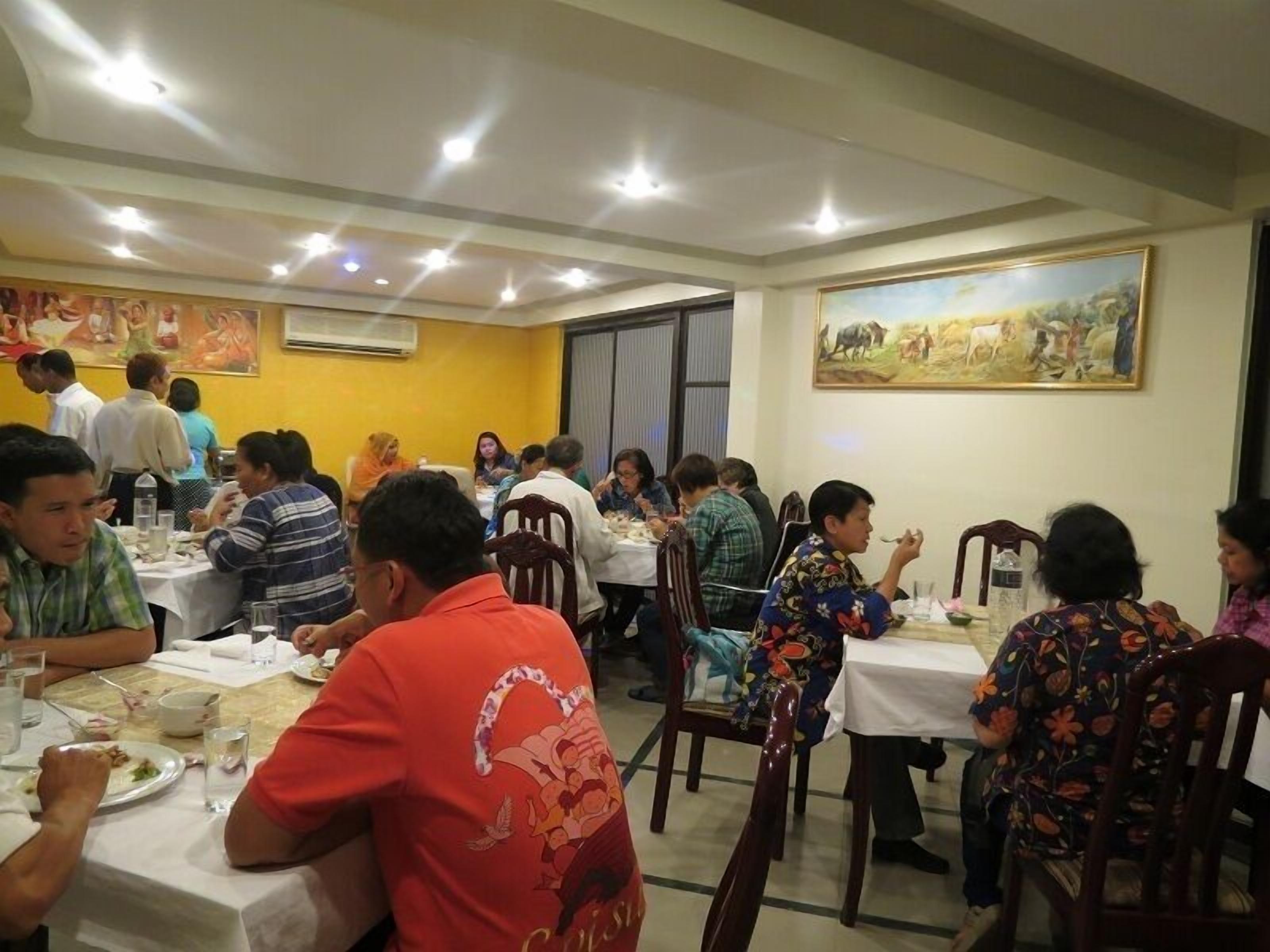 A restaurant with multiple tables and chairs, people dining, and a painting on the wall.