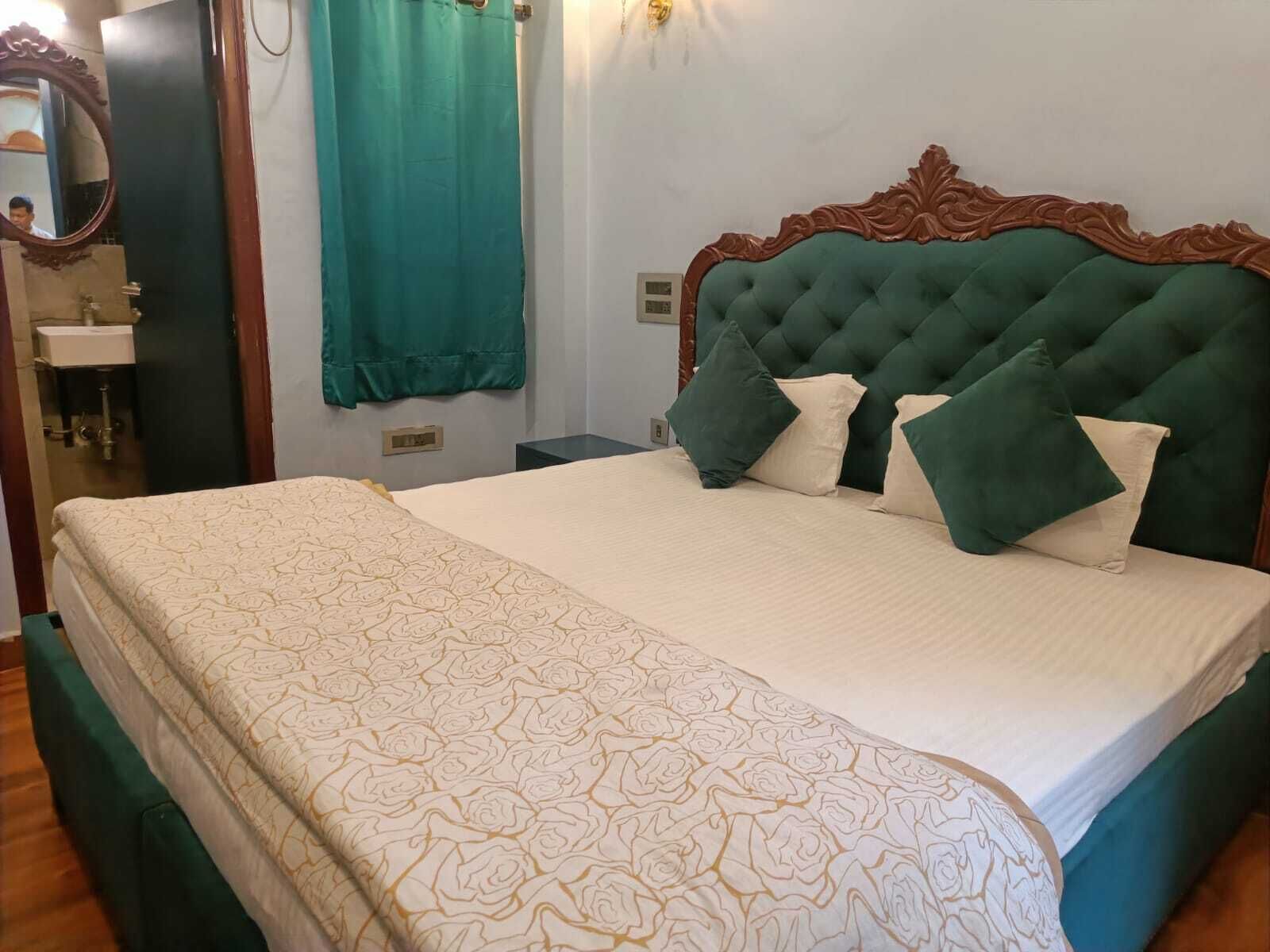 A neatly made bed with a green headboard and multiple pillows, a bathroom with a mirror and sink, and a teal curtain.