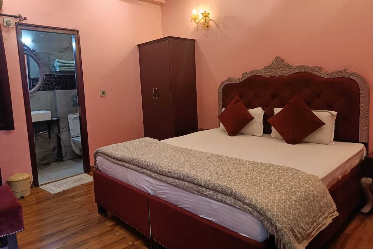 A bedroom with a large bed, a wardrobe, a bathroom visible through an open door, and a wall-mounted light fixture.
