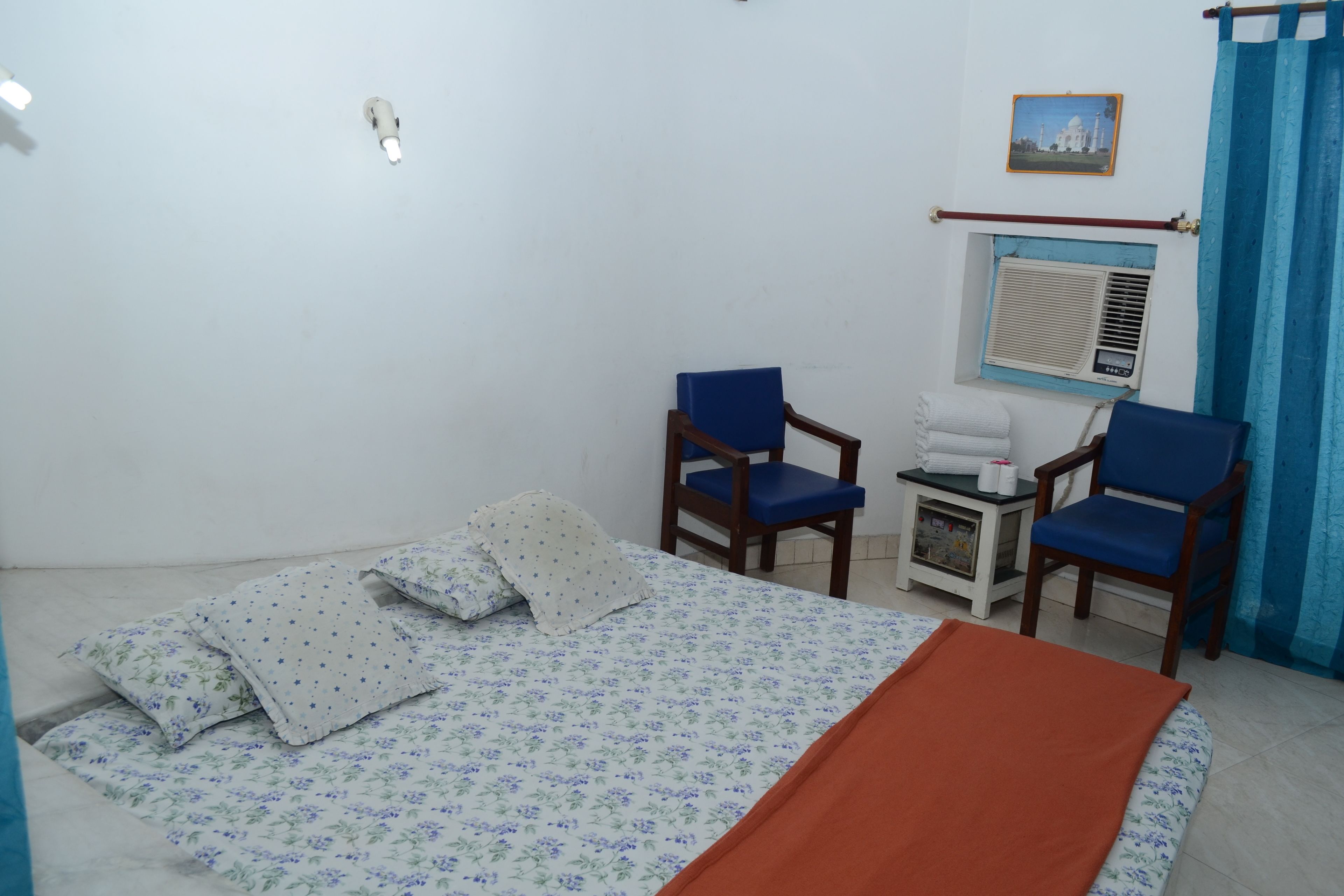 Una habitación con una cama, dos sillas azules, una mesita con mantel blanco, una ventana con cortina azul y un cuadro en la pared.