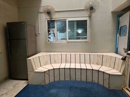 A compact kitchen with a built-in curved bench, two wall-mounted fans, a window, and a refrigerator.