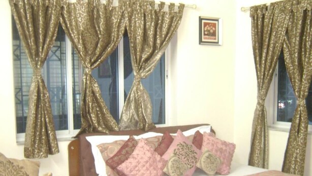 A bedroom with two beds, patterned curtains, a bedside table with a vase of flowers, and a framed picture on the wall. - Innra (Kolkata)