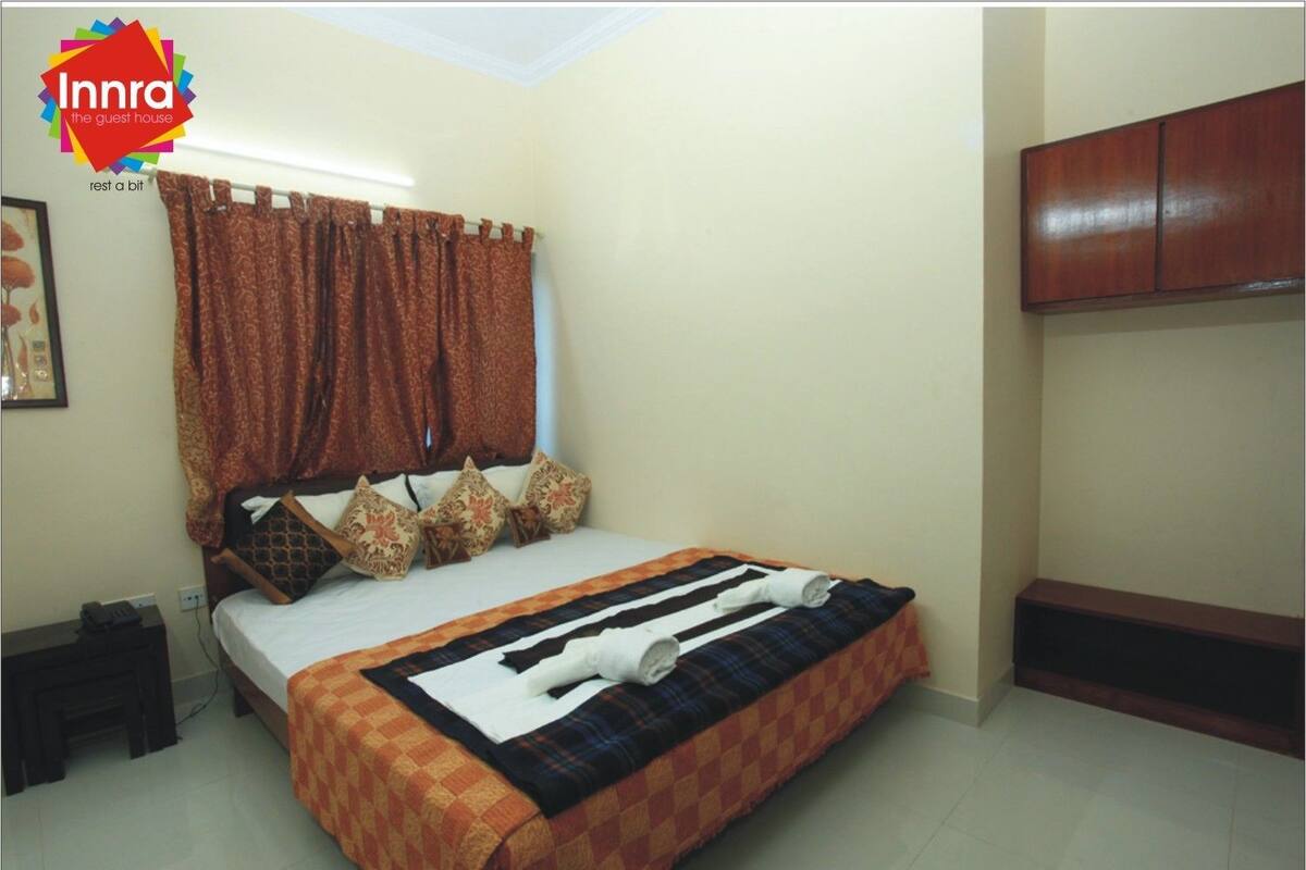 A neatly made bed with patterned pillows and a striped blanket, a wooden wardrobe, and a small bedside table with a phone.