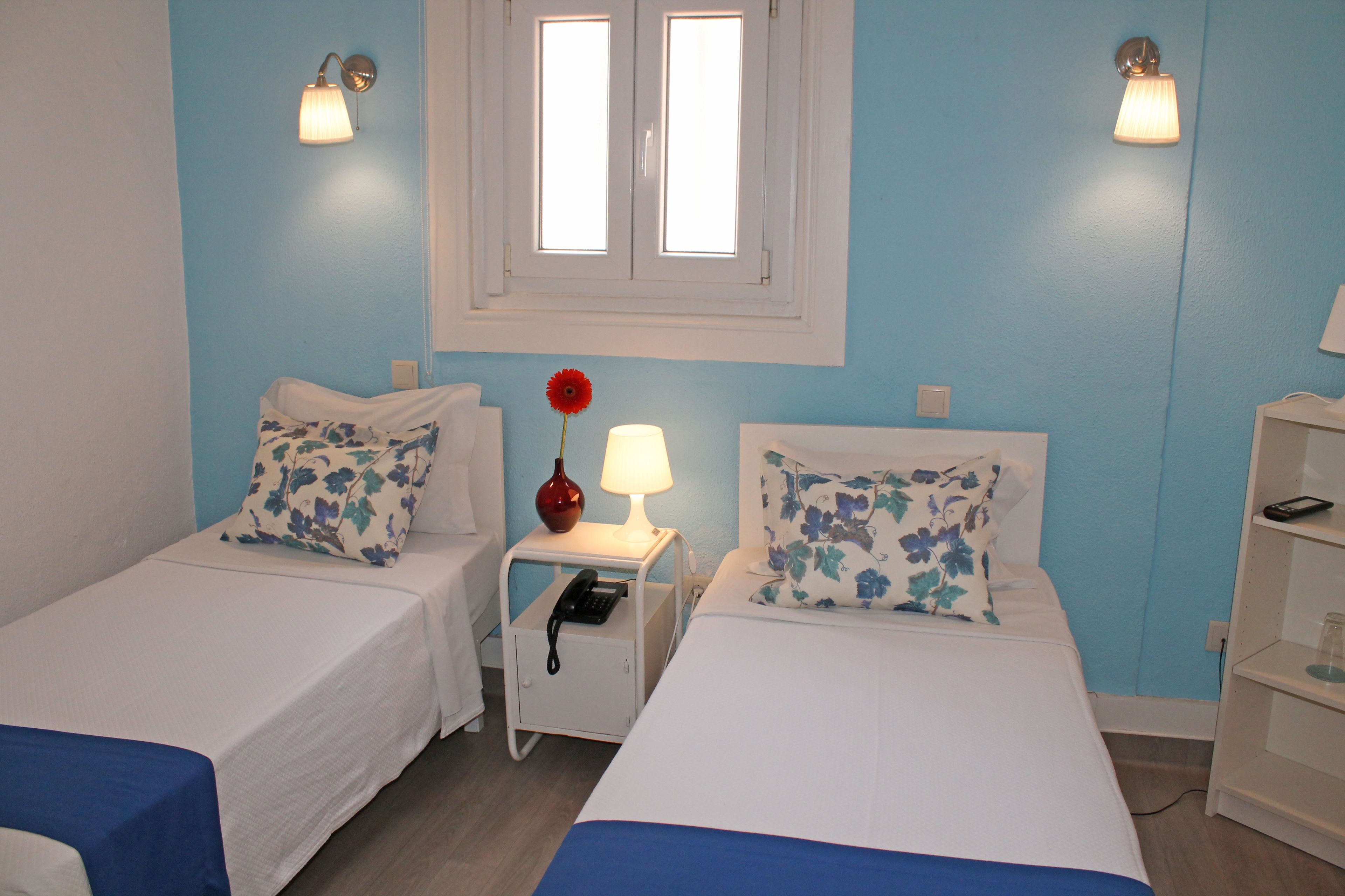 Habitación de hotel con dos camas, cada una con almohada floral, una mesita de noche con lámpara y un jarrón rojo.