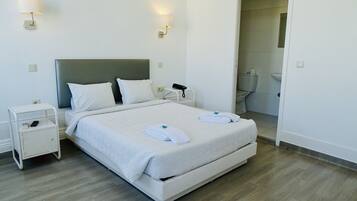 Una cama bien hecha con sábanas blancas y dos toallas dobladas, una mesita de noche con un teléfono y un control remoto, y una lámpara fijada a la pared.