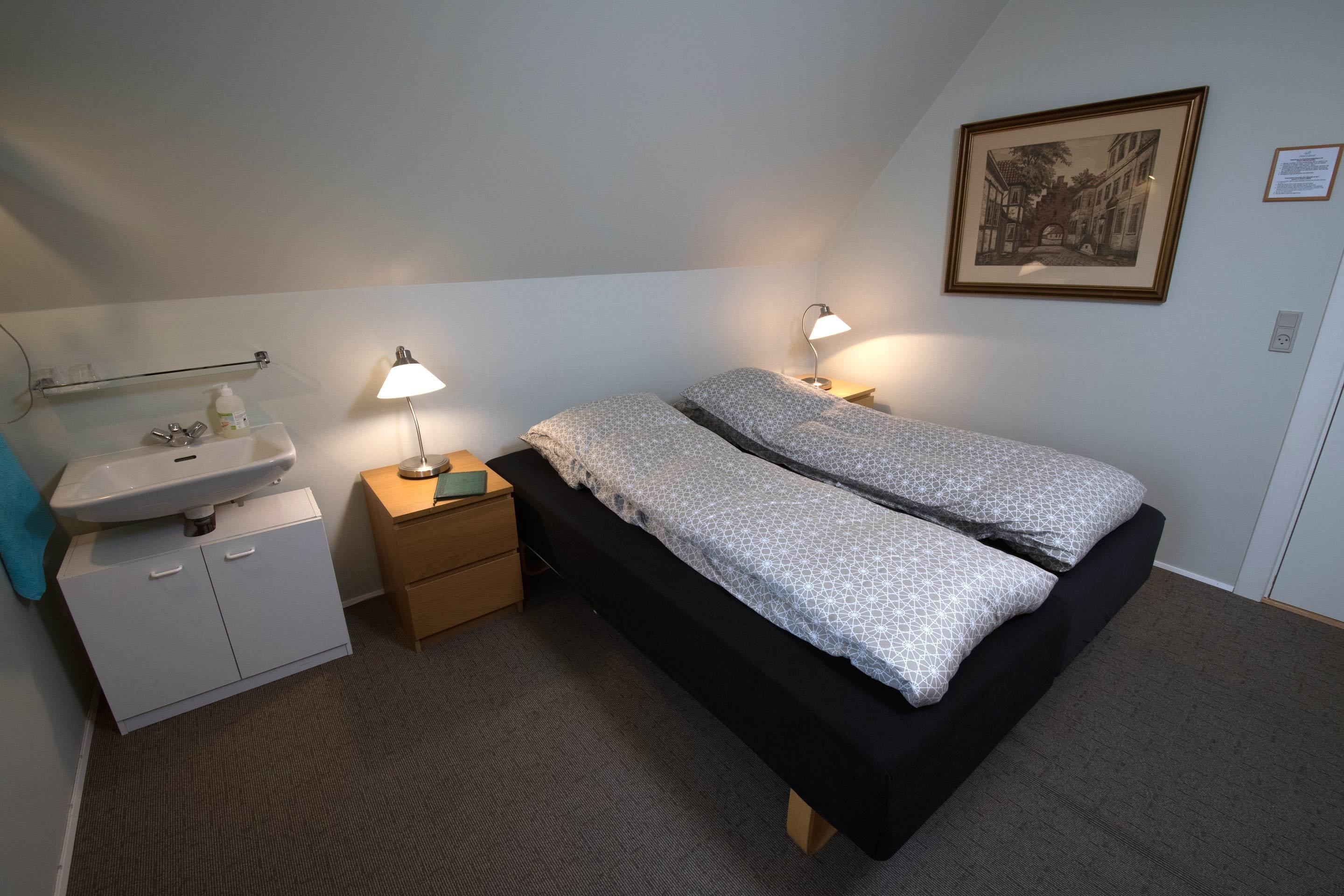 Un dormitorio con cama, lámparas de noche, una mesita de noche, un lavamanos instalado en la pared y un cuadro enmarcado.