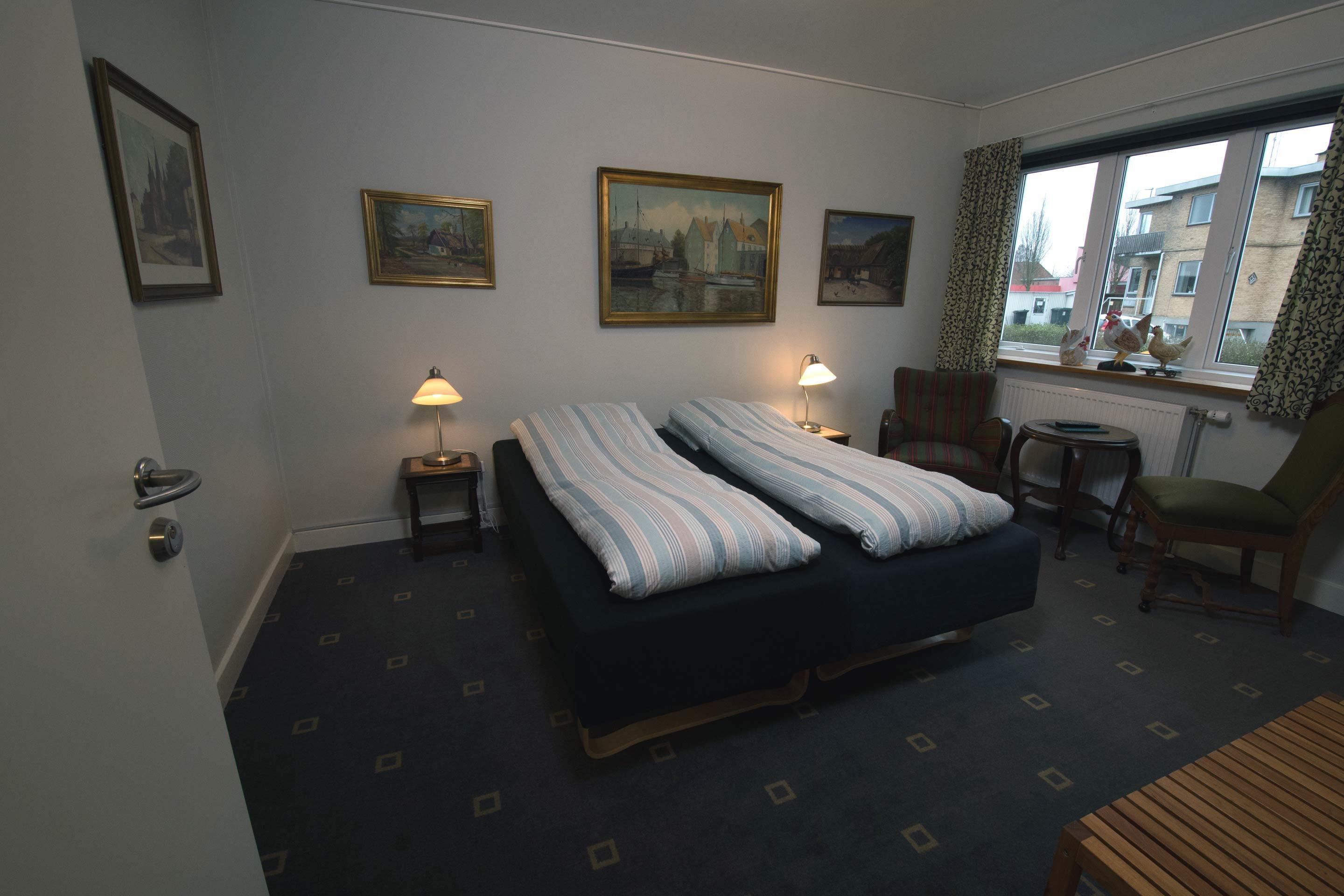 A hotel room with two beds, a chair, a small table, and a window with a view of buildings.