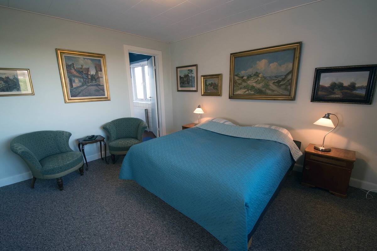 A bedroom with a large bed, two armchairs, a nightstand with a lamp, and several framed paintings on the walls.