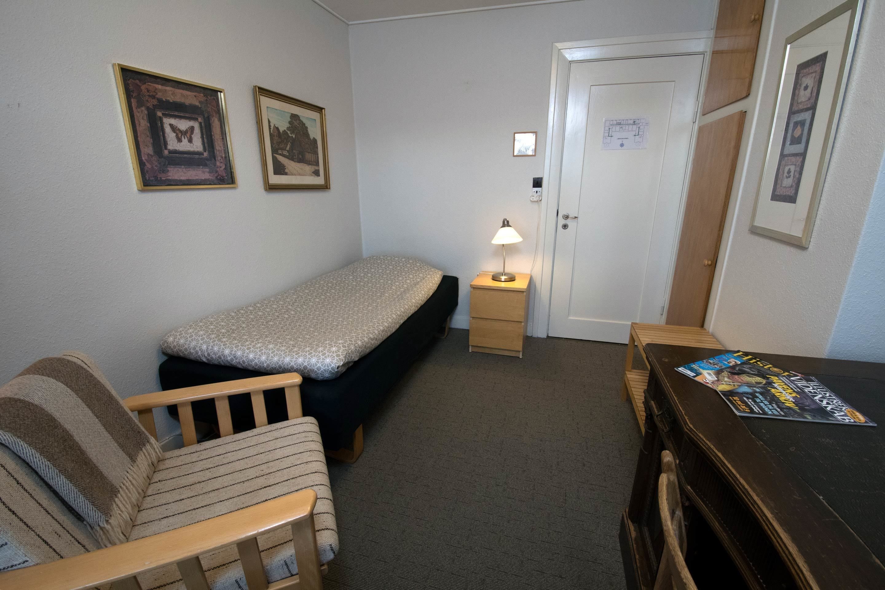 A small, neatly arranged room with a single bed, a wooden chair, a nightstand with a lamp, and a desk with a magazine.