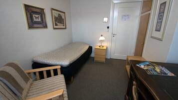 A small, neatly arranged room with a single bed, a wooden chair, a nightstand with a lamp, and a desk with a magazine.