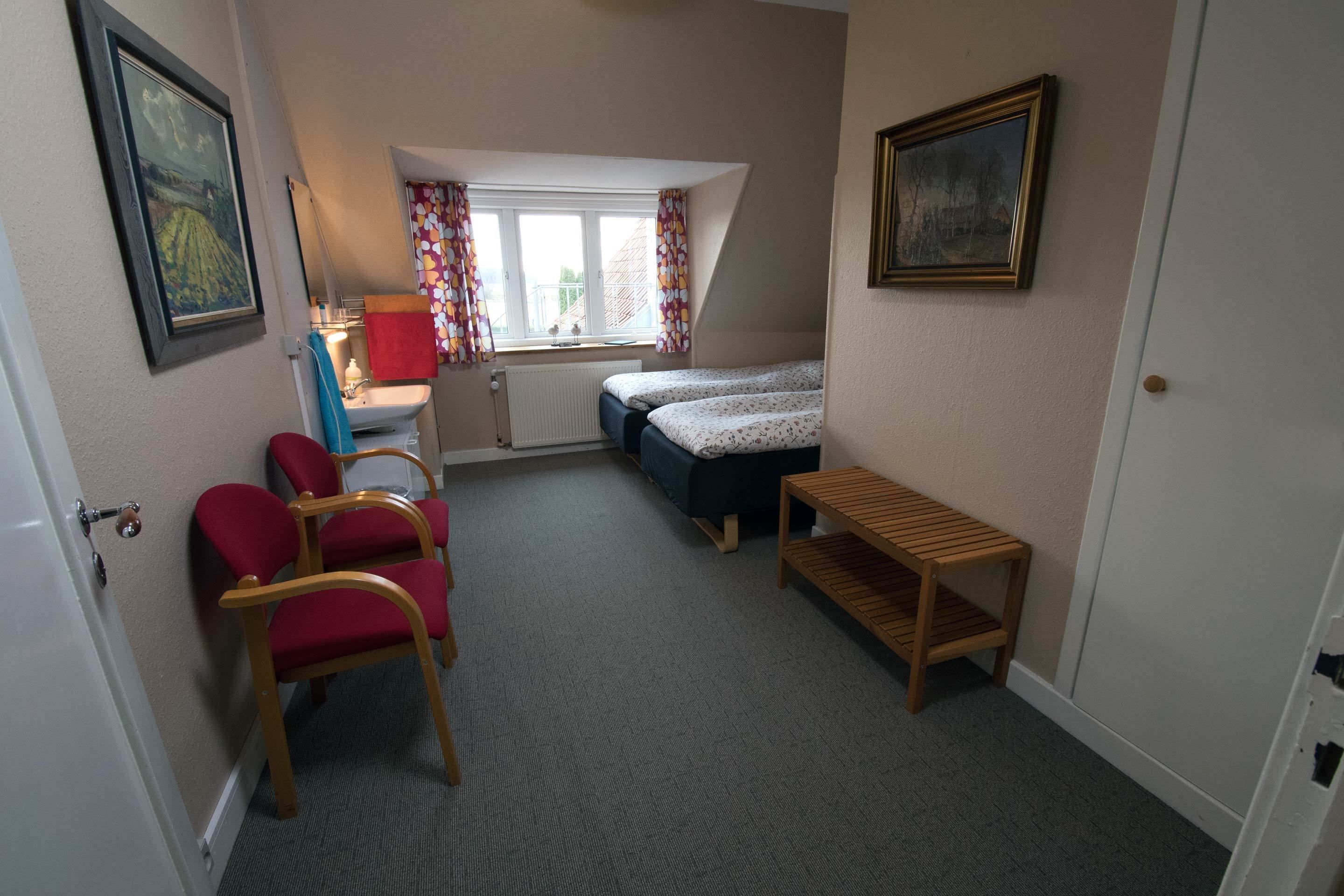 A small, well-lit room with a bed, a desk, and a chair. There is a window with curtains, a radiator, and a framed picture on the wall.