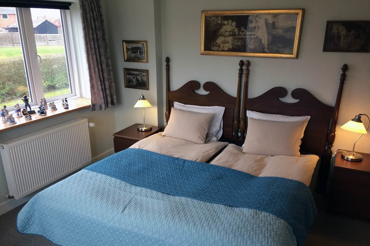 A bedroom with a large bed, two bedside tables with lamps, a window with curtains, and a radiator.