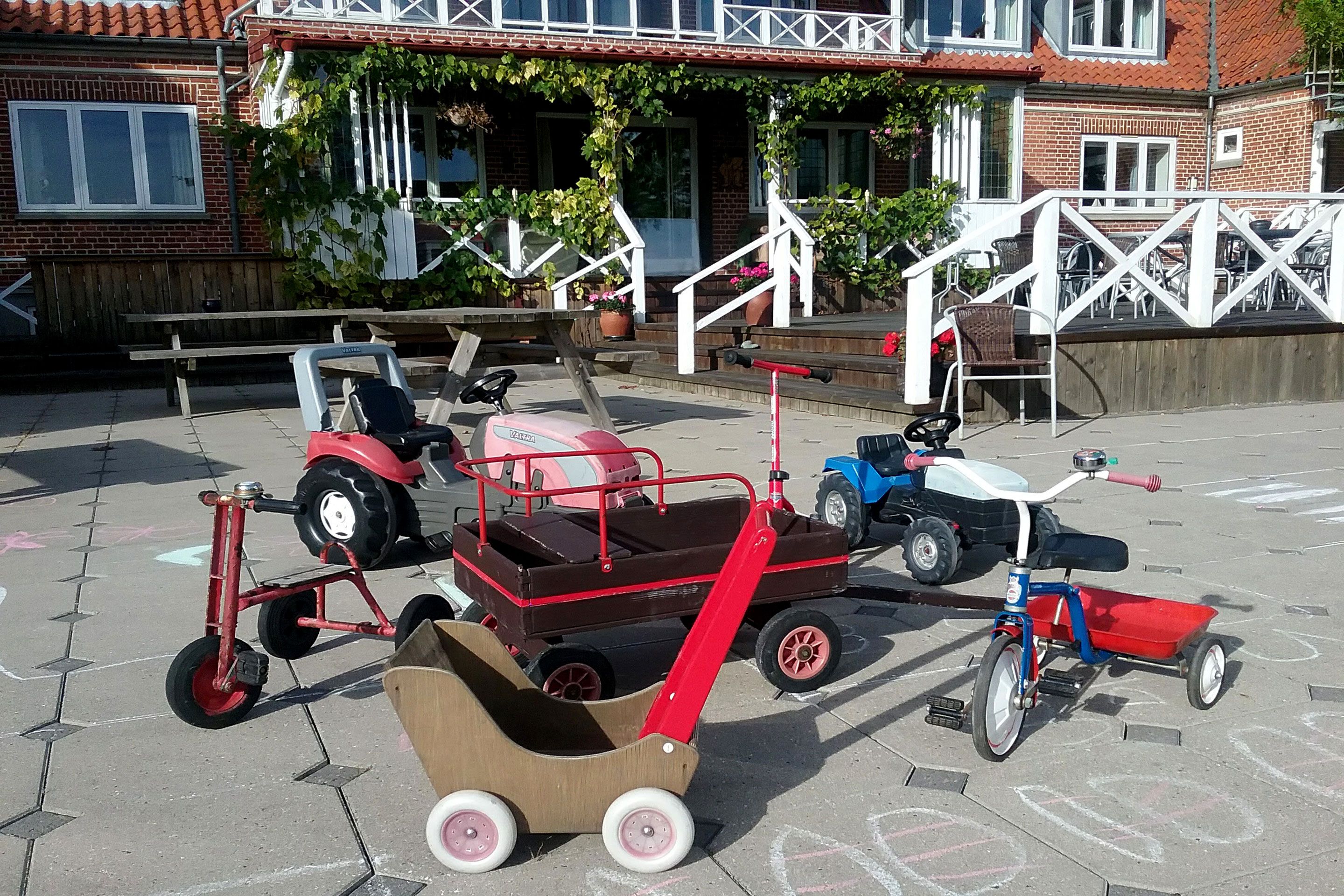 A patio area with a wooden bench, a red tricycle, a blue tricycle, and a small wooden cart.