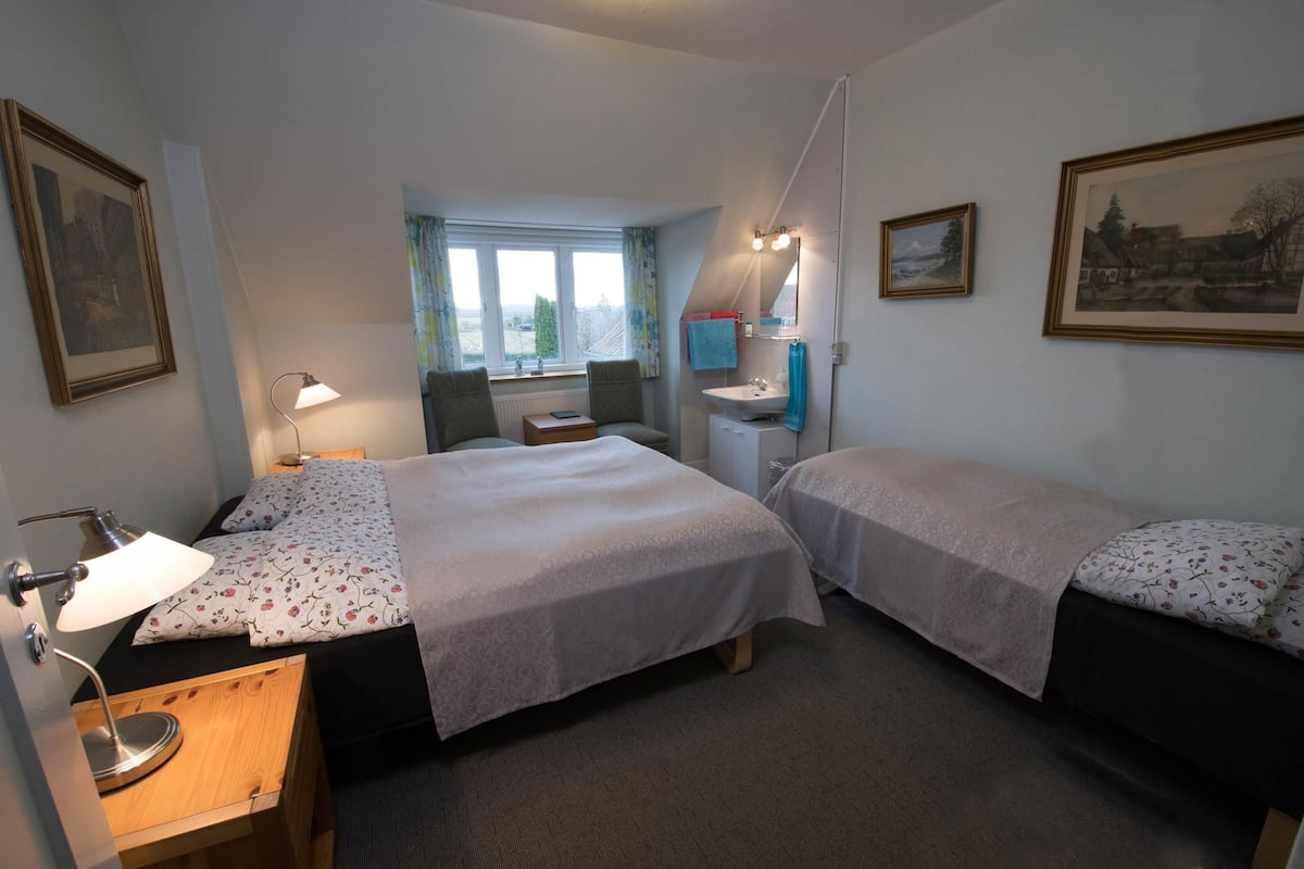 A hotel room with two beds, a desk with a lamp, a window with curtains, a sink, and framed pictures on the walls.