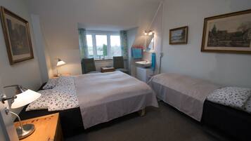 A hotel room with two beds, a desk with a lamp, a window with curtains, a sink, and framed pictures on the walls.