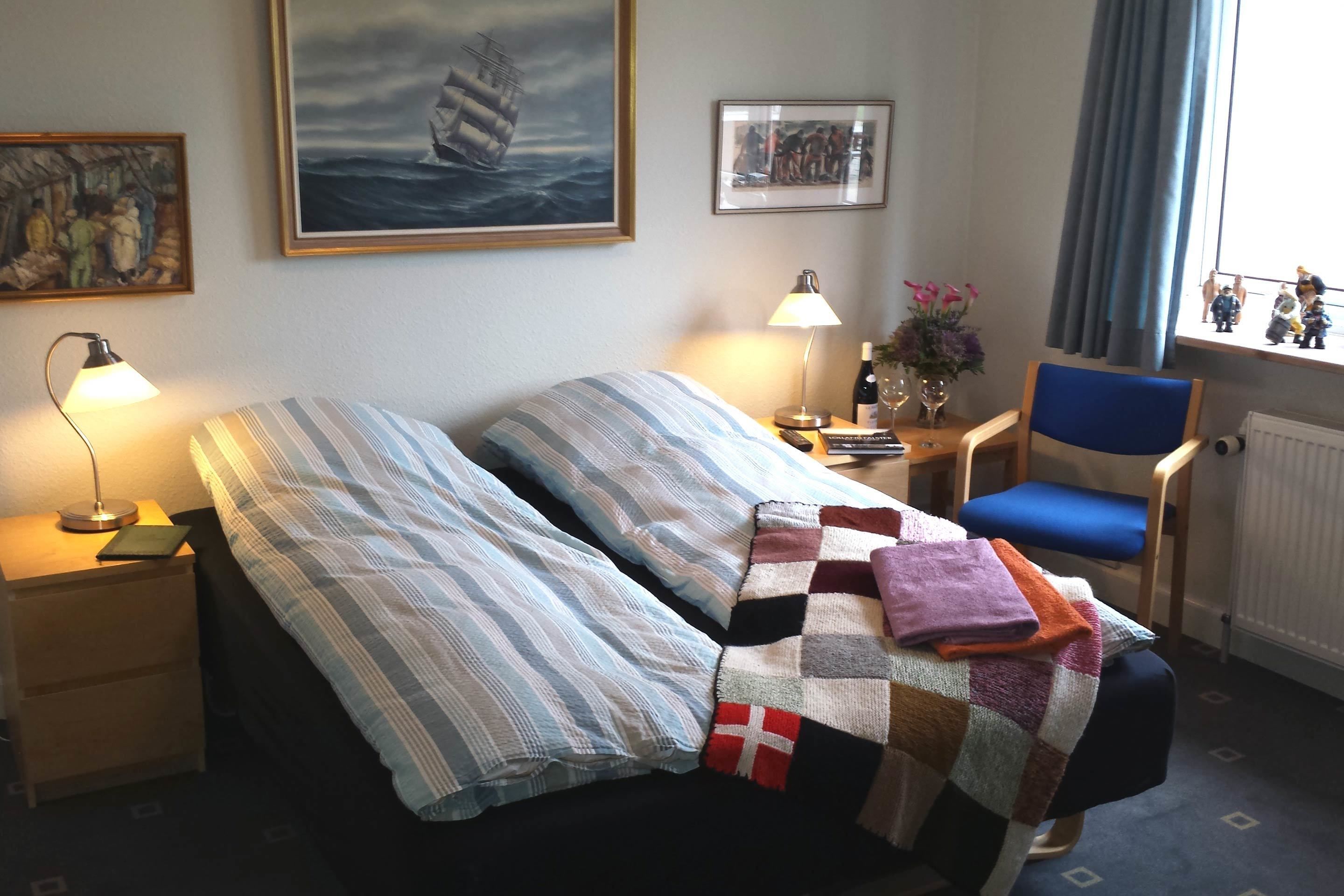 Une chambre d’hôtel avec un lit, deux lampes de chevet, une chaise, une petite table ornée d’un vase de fleurs et des œuvres d’art accrochées aux murs.