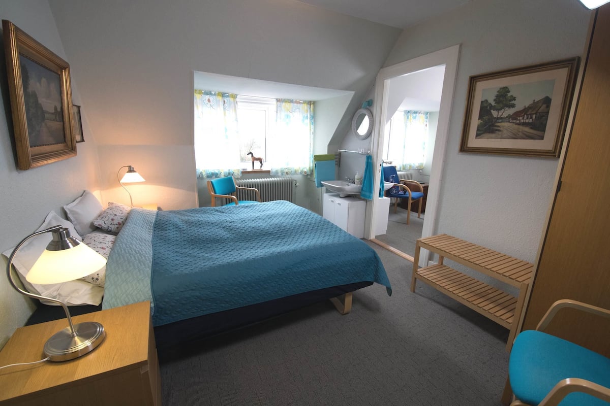 A hotel room with a bed, a desk with a lamp, a chair, a small table, and a window with curtains.