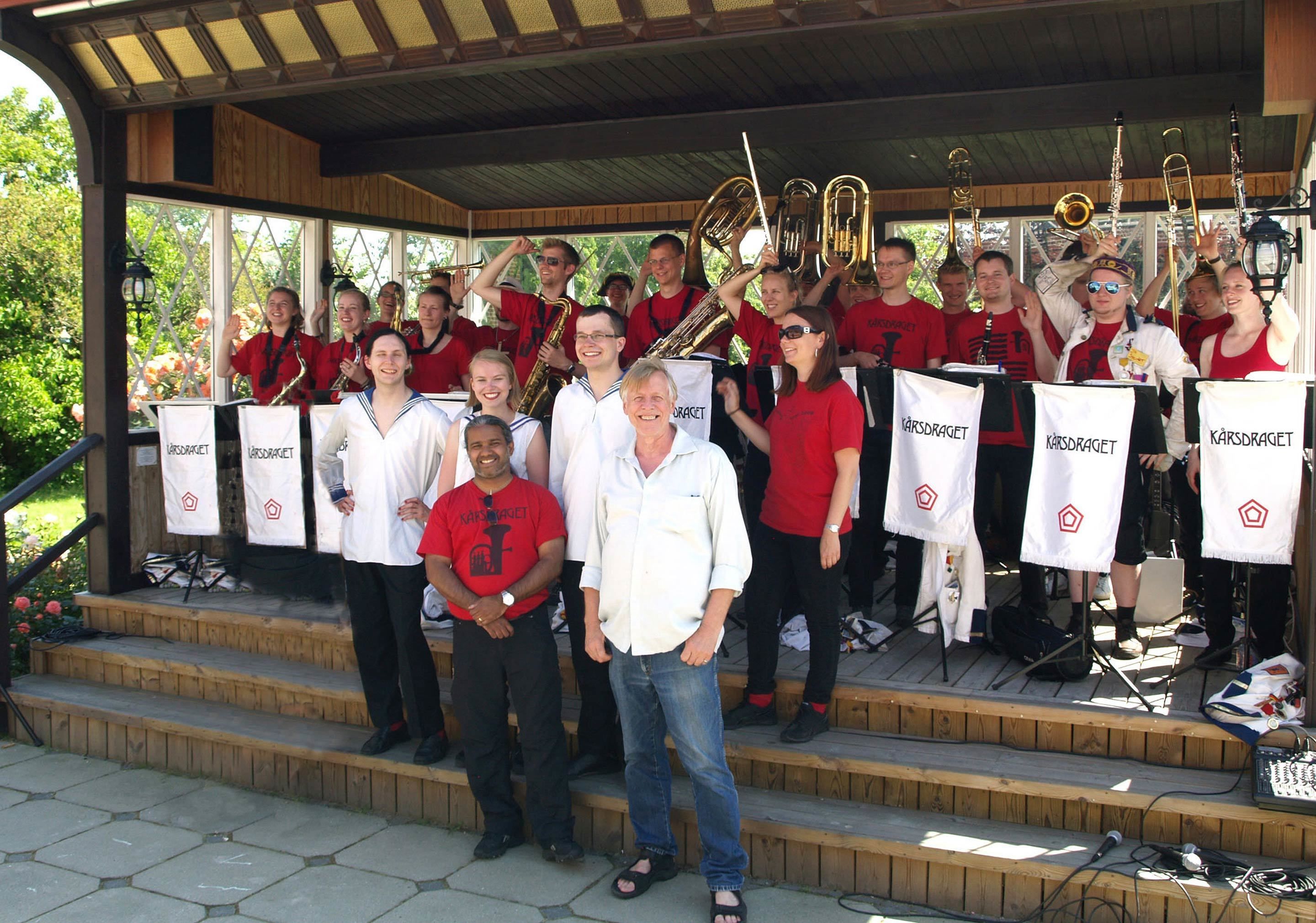 Un grupo de personas en un escenario con instrumentos musicales, vestidas con uniformes rojos y blancos a juego, bajo una estructura cubierta con escalones de acceso.