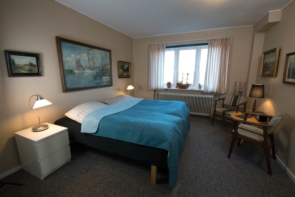 A bedroom with a bed, bedside tables, a desk, chairs, a window with curtains, and wall art.