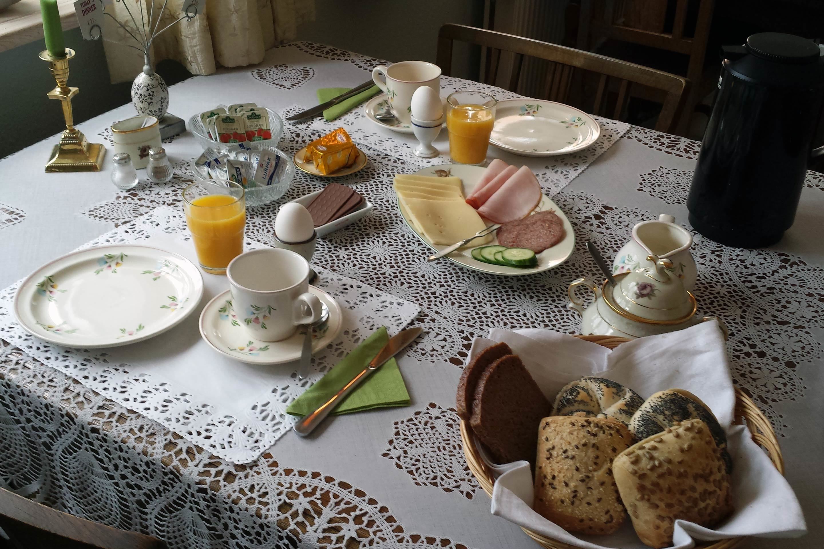 A breakfast table set with plates, cups, a teapot, a thermos, and a basket of bread.