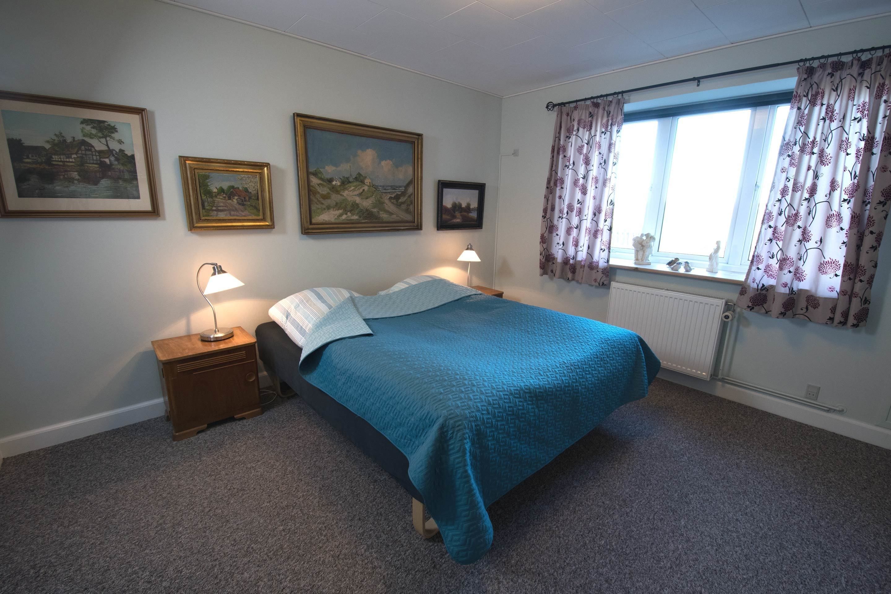 Ein Schlafzimmer mit einem Bett, zwei Nachttischlampen, einem Fenster mit floralem Vorhang und drei gerahmten Bildern an der Wand.