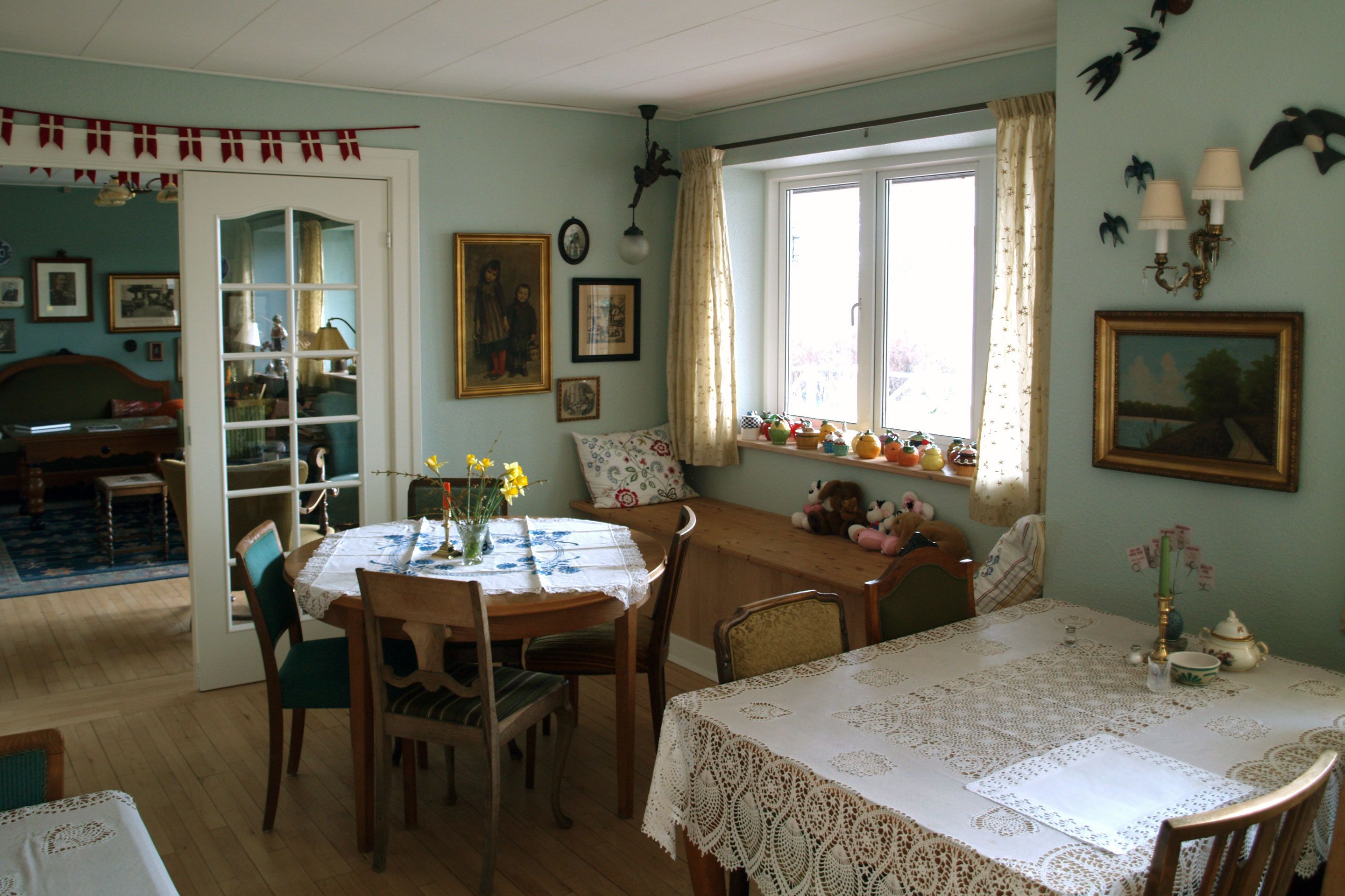 Un coin repas accueillant, avec une table dressée pour quatre personnes, entourée de chaises en bois. On aperçoit, à l’arrière-plan, une bibliothèque remplie de livres et d’objets décoratifs.